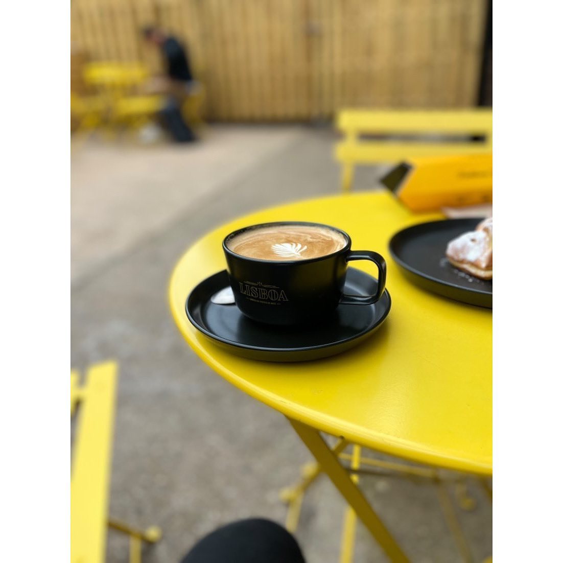 A cappuccino sit on a bright yellow metal table, outside.