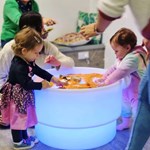 Two young children playing at a round, illuminated sensory table filled with orange material. They are using their hands to explore the contents, with an adult nearby supervising. The setting appears to be an indoor play or educational area with cushions and furniture in the background.