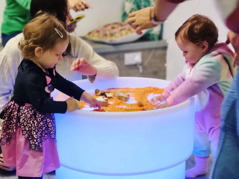 Two young children playing at a round, illuminated sensory table filled with orange material. They are using their hands to explore the contents, with an adult nearby supervising. The setting appears to be an indoor play or educational area with cushions and furniture in the background.