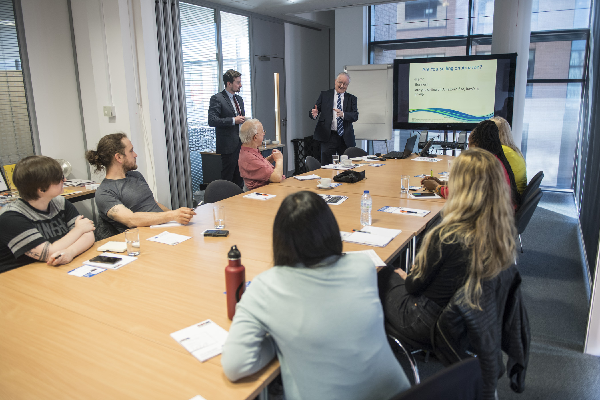 Workshop attendees sit round a large table in a modern office watching a presentation. 