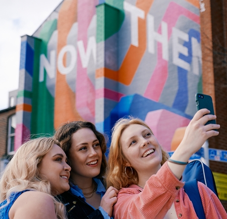 Three women take a selfie in front of the Now Then mural on Howard Street in Sheffield city centre.