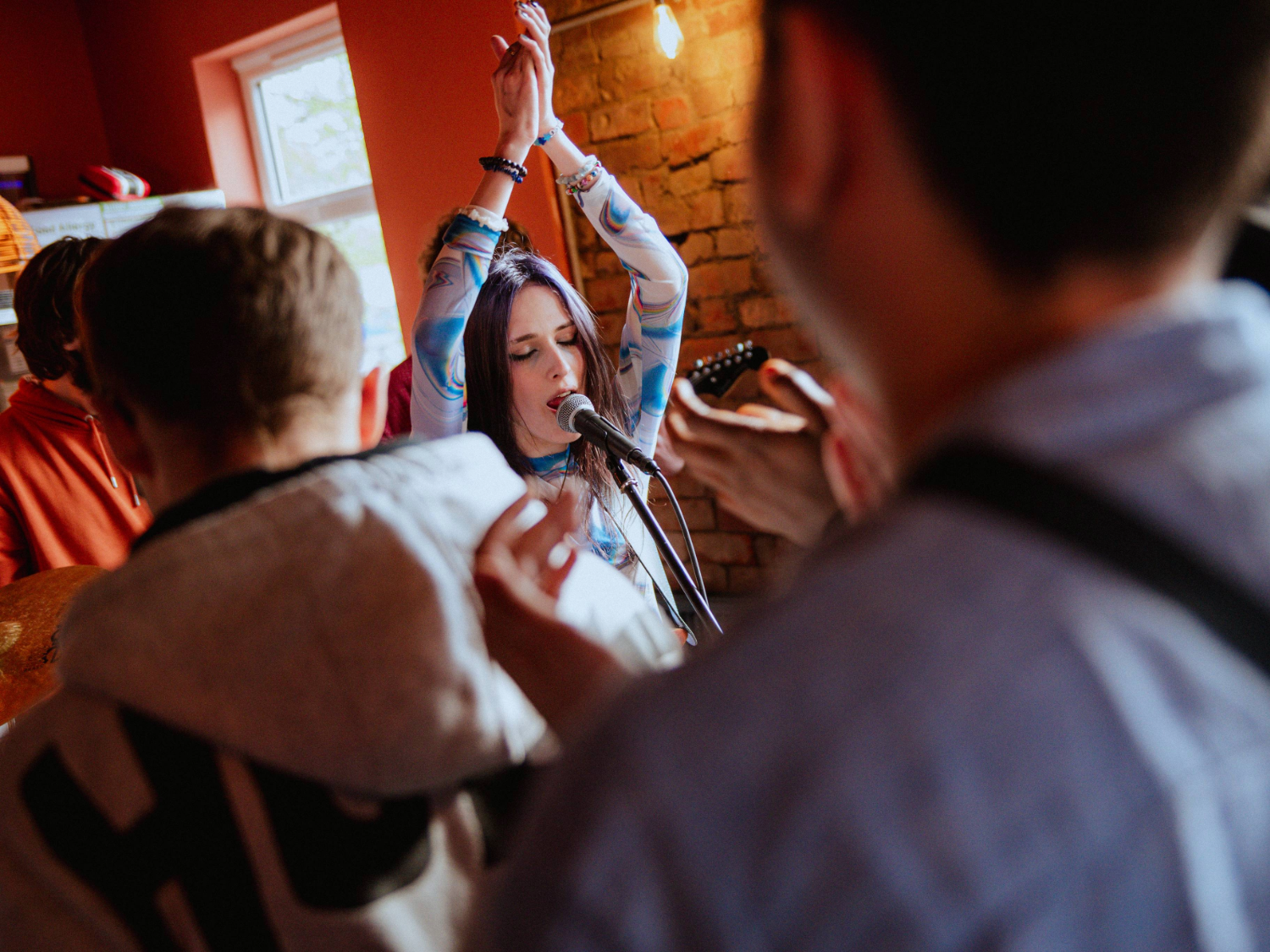 Indoor scene with exposed brick wall and warm lighting. A person stands in front of a microphone with arms raised, wearing a patterned long-sleeve top. Several people are gathered around, partially visible in the foreground, creating a lively atmosphere.