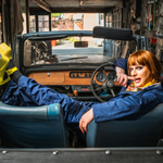 A man, in make-up and a wig and wearing blue overalls and bright yellow platform shoes, lounges in an open top-car in a small garage that is full of all sorts of tools, spare-parts and cloths and rags.