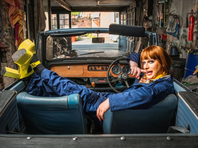 A man, in make-up and a wig and wearing blue overalls and bright yellow platform shoes, lounges in an open top-car in a small garage that is full of all sorts of tools, spare-parts and cloths and rags.