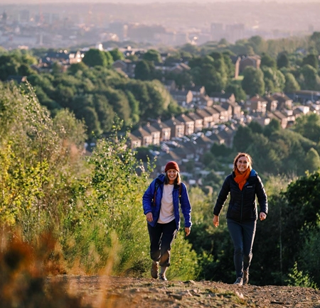 Two ramblers are walking up a hill. Down in the valley below them you can see rows of terrace houses and on the horizon you can see Sheffield city centre.