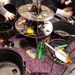 Halloween-themed sensory play setup with a black multi-tiered stand covered in spider webs and plastic spiders. Surrounding it are black cauldrons and bowls filled with toy insects, bats, and pumpkins. The background features purple rice or sand, enhancing the spooky atmosphere.
