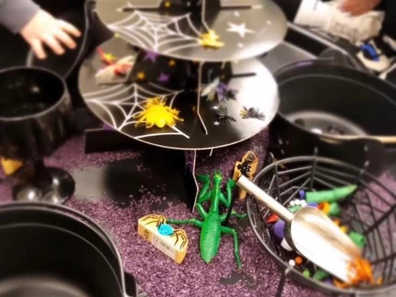 Halloween-themed sensory play setup with a black multi-tiered stand covered in spider webs and plastic spiders. Surrounding it are black cauldrons and bowls filled with toy insects, bats, and pumpkins. The background features purple rice or sand, enhancing the spooky atmosphere.