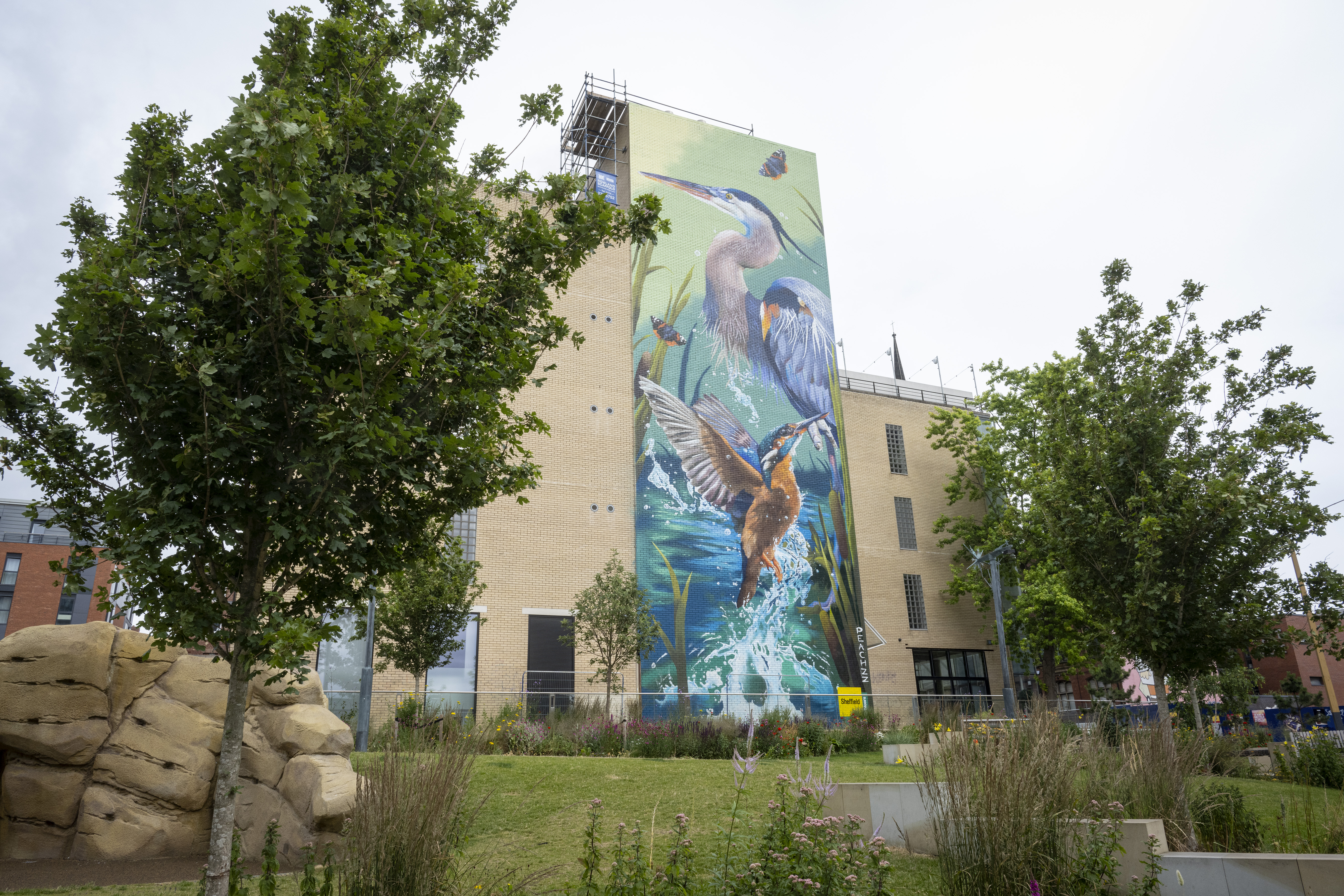 A tall beige building with a large, colorful mural depicting birds in flight and aquatic plants. The mural features a heron and a kingfisher against a vibrant blue and green background. In the foreground, there are trees, landscaped grass, flowers, and large rock formations, creating a natural park-like setting under a cloudy sky.
