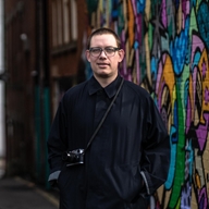 A man in a long, dark coat, with a camera slung over his shoulder, stands in front of some colourful street art.
