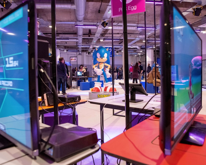Interior of the National Videogame Museum with multiple gaming monitors in the foreground and a large Sonic the Hedgehog figure in the center. People are scattered around the space, interacting with exhibits. The room has an industrial ceiling with exposed pipes and bright lighting, creating a modern gaming environment.