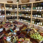 Rows of shelving filled with bottles of wine. In the centre of the room is a table covered in artfully arranged cheese, meats, grapes and other goods for sale.