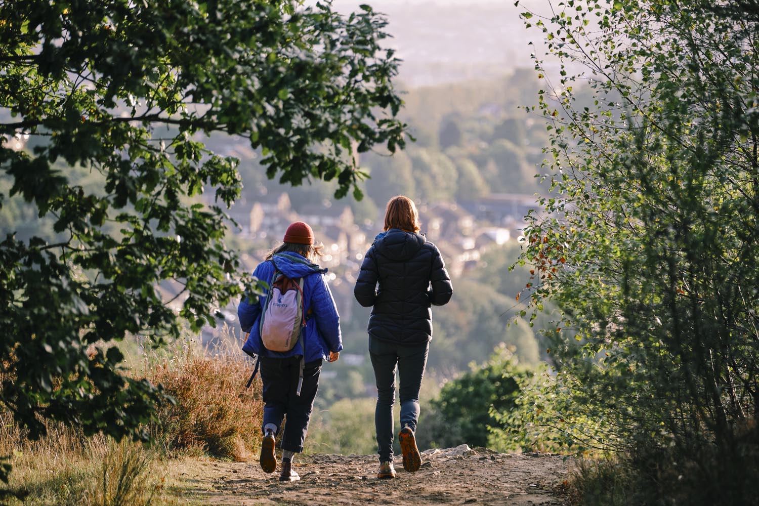 Two people walking along a dirt trail through a wooded area, wearing outdoor jackets and backpacks. The path leads downhill toward a scenic view of a town nestled among trees in the background. Sunlight creates a warm glow over the distant landscape, with green foliage framing the scene.