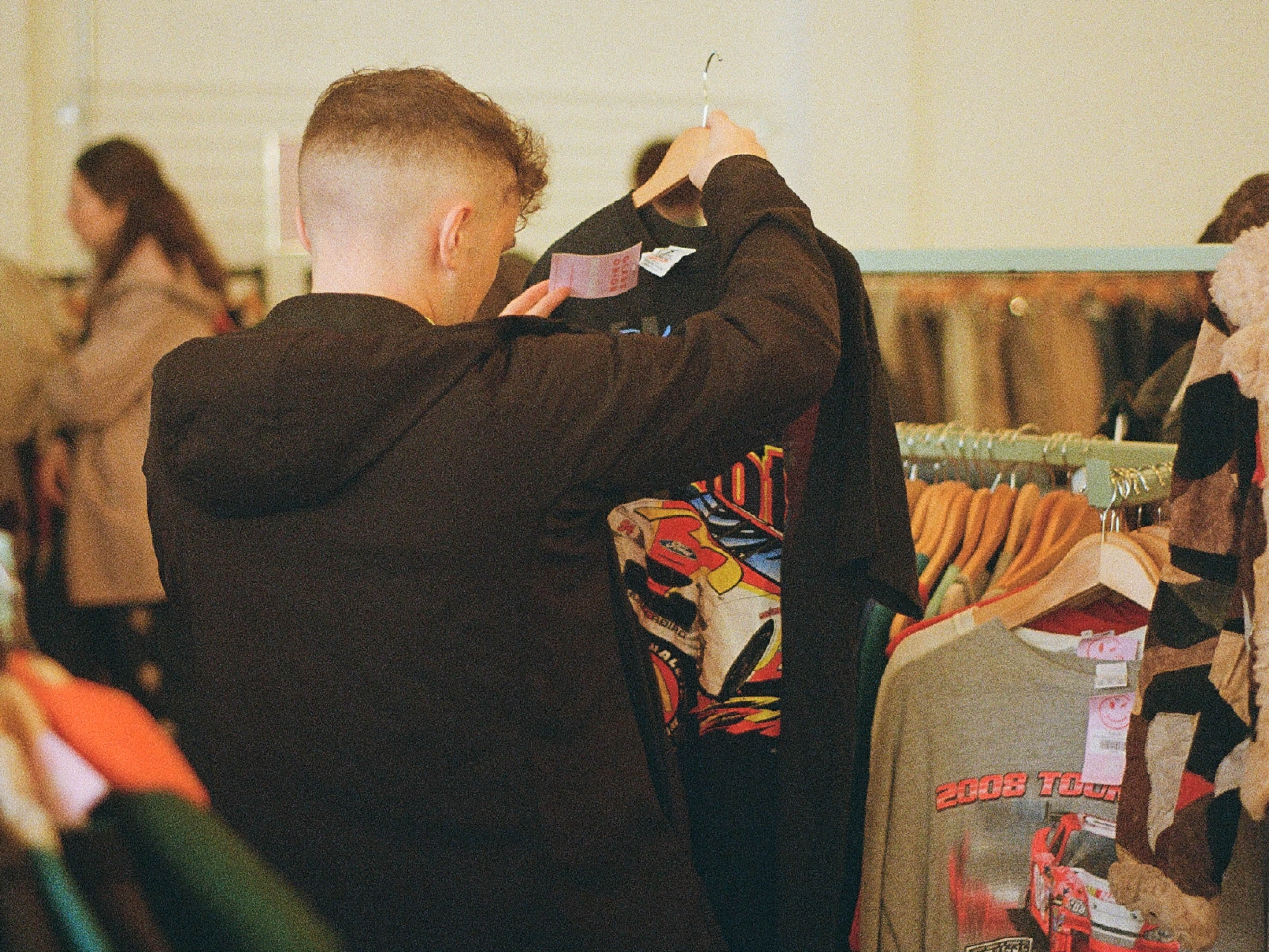 A person wearing a dark jacket is browsing through clothing on a rack in what appears to be a vintage or second-hand store. The individual is holding up a black T-shirt featuring a colorful graphic design, while other shirts with printed designs hang nearby. The background shows more clothing racks and shoppers in the store.
