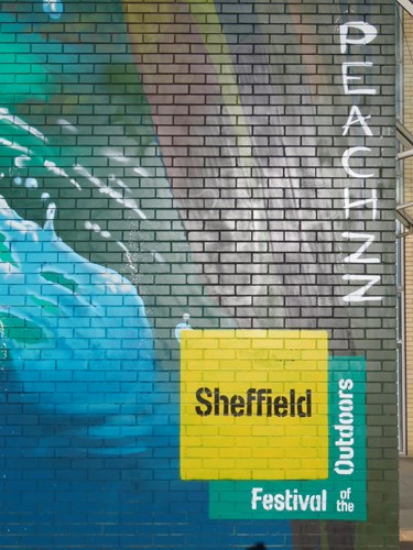 Close-up of a painted brick wall featuring part of a mural with blue and green water-like patterns. The word ‘PEACHZZ’ is written vertically in white letters on the right side. At the bottom, a yellow and green block of text reads ‘Sheffield Festival of the Outdoors .’