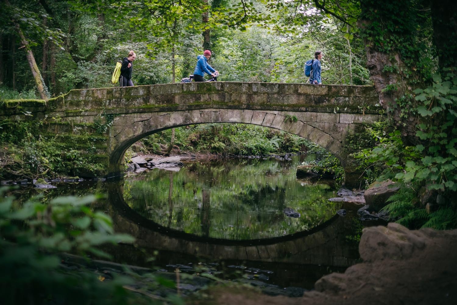 Three people walking across an old stone bridge in a lush forest setting. The bridge arches over a calm stream that reflects the greenery and the shape of the arch, creating a near-perfect circle in the water. The surrounding area is dense with trees, vines, and undergrowth, giving the scene a natural and tranquil atmosphere.
