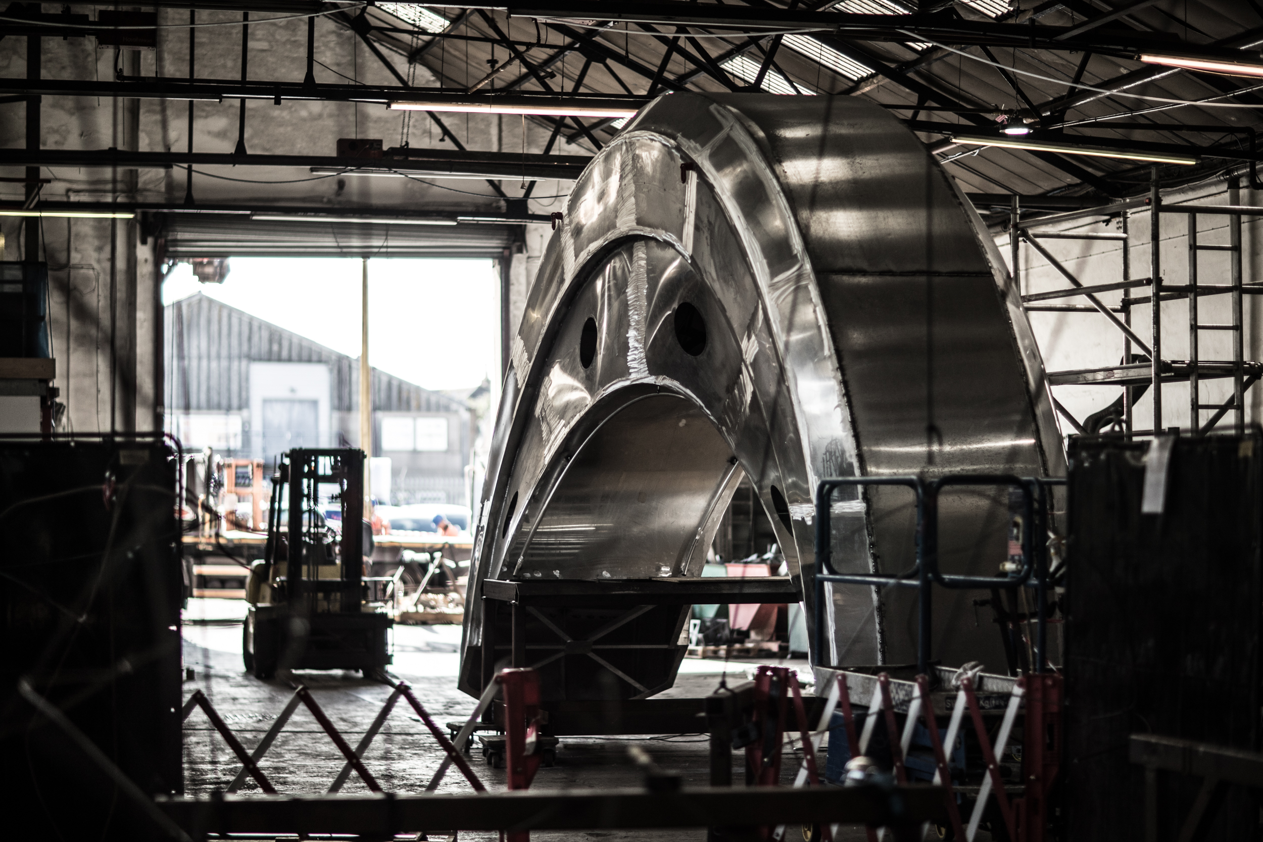 Large metallic structure resembling a curved boat hull under construction inside an industrial workshop. The piece is shiny and partially assembled, with scaffolding and barriers around it. A forklift and open workshop doors are visible in the background, along with steel beams and equipment.