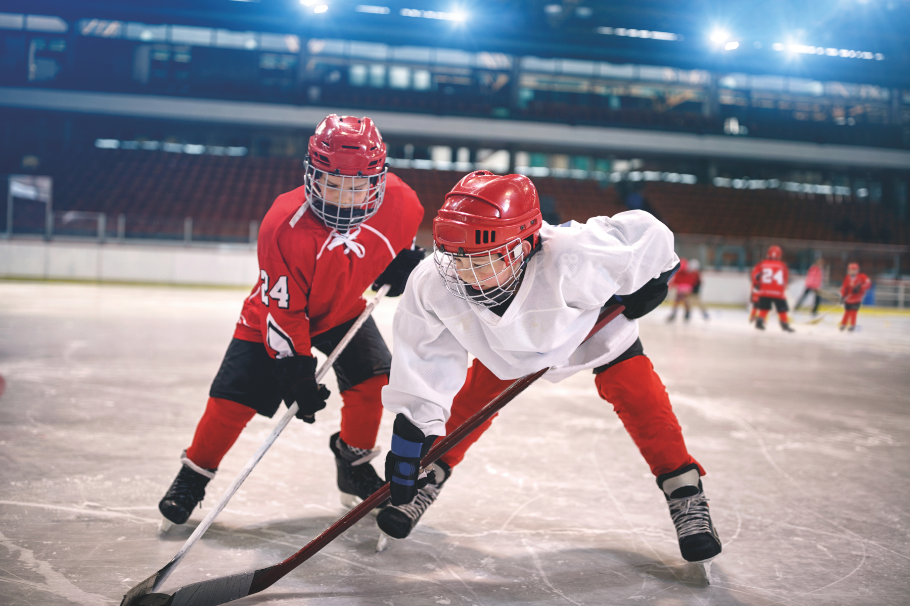Two ice hockey players in full gear competing for the puck on an indoor rink. One player wears a white jersey and red pants, leaning forward with a stick on the ice, while the other in a red jersey closely defends. Additional players and seating are visible in the background under bright arena lights.
