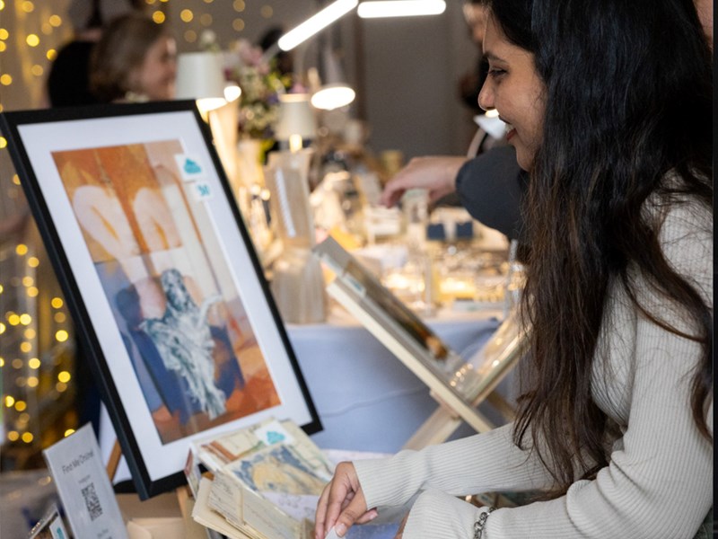 A person views a framed abstract artwork at an art or craft fair. The artwork features warm tones with blue accents. Surrounding the display are books, decorative items, and string lights, creating a cozy and creative atmosphere.