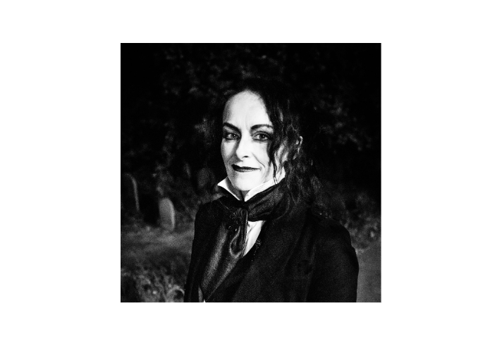 Rosie Garland: A black and white photograph of a person with long, wavy hair, dressed in a dark suit with a high-collared white shirt and cravat. They wear dark eye makeup and lipstick, contributing to a gothic aesthetic. The dimly lit background features gravestones and foliage, enhancing the moody, atmospheric tone of the image.