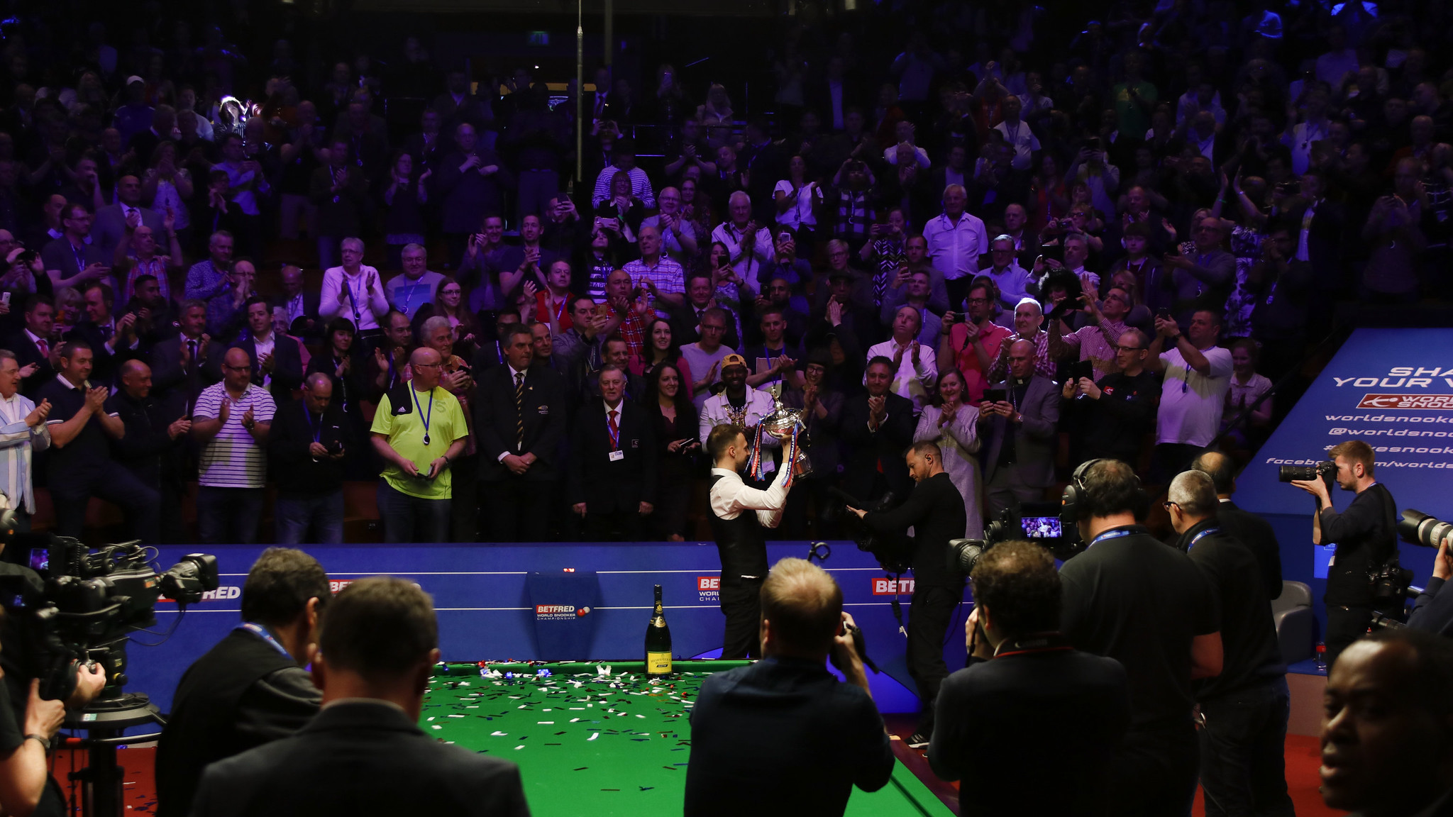 Snooker championship celebration at an indoor arena. A player stands at the snooker table holding a trophy while photographers capture the moment. Green table surface is covered with confetti and a champagne bottle is placed on it. Behind the table, a large crowd of spectators applauds under bright stage lighting.