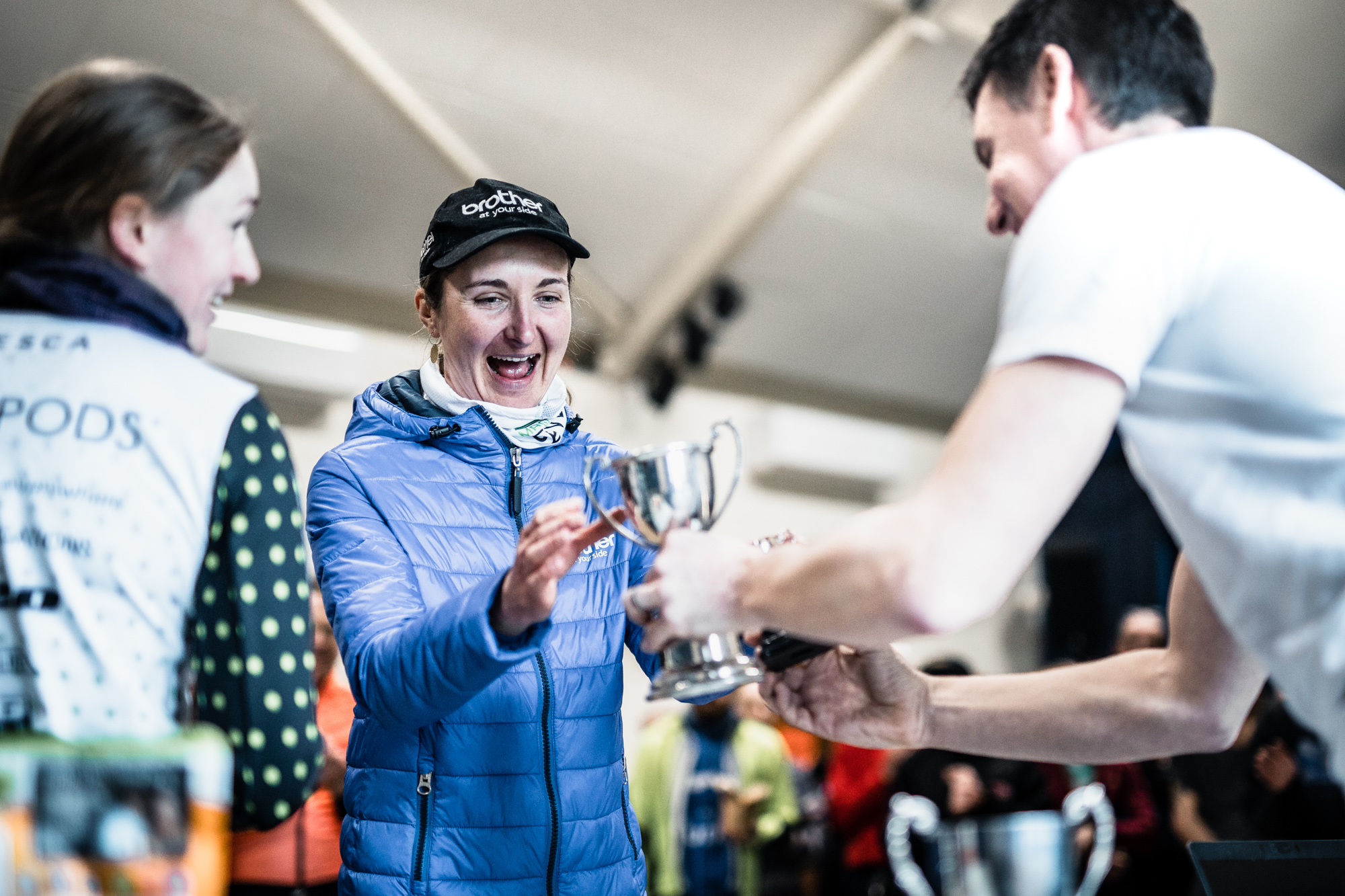 Woman excited as she receives a small silver trophy being handed to her by a man. Another female is also in conversation to the left.