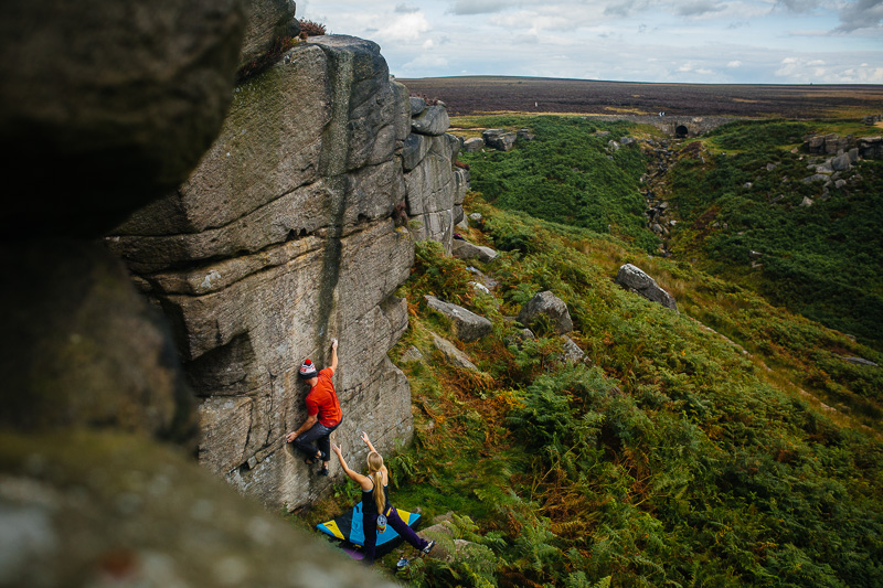 Two people are climbing up a rock face. They are using climbing gear and have a safety mat on the ground.