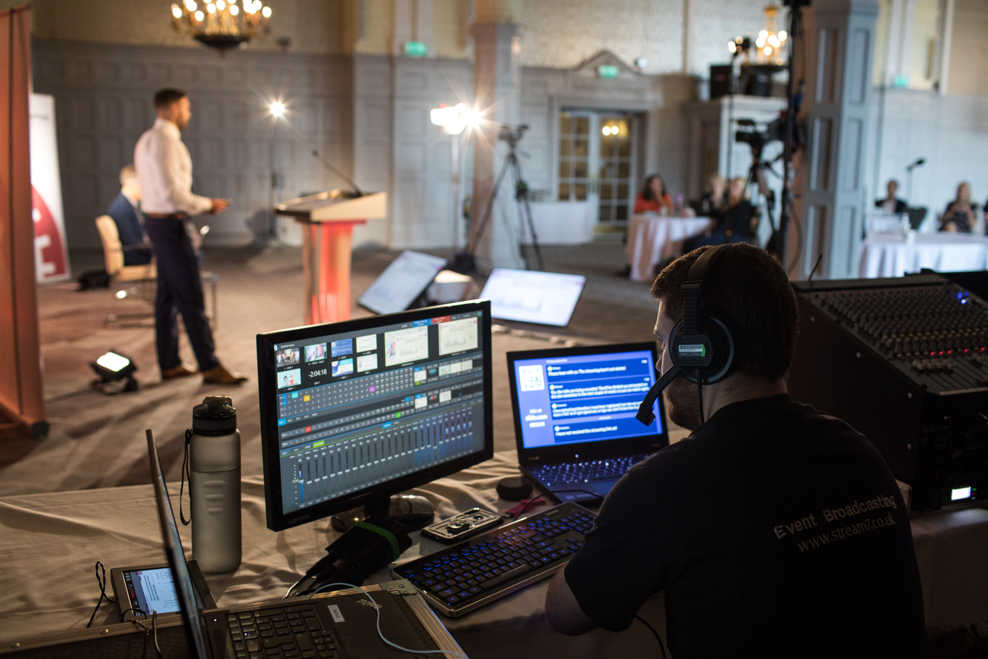 A live event production setup showing a technician operating multiple screens and audio equipment in the foreground, while a speaker stands at a podium in the background. The room is equipped with cameras, lighting, and monitors for broadcasting.
