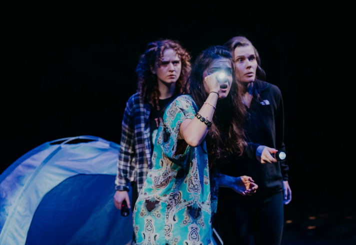 Three women, onstage as part of a play, are looking worried. Two of them are holding torches. Behind them is a blue tent.