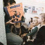 A person reads the children's book "Room on the Broom" by Julia Donaldson and Axel Scheffler to a group of young children seated attentively. The book cover shows a witch flying on a broomstick with animals. The background includes a blurred classroom with posters.