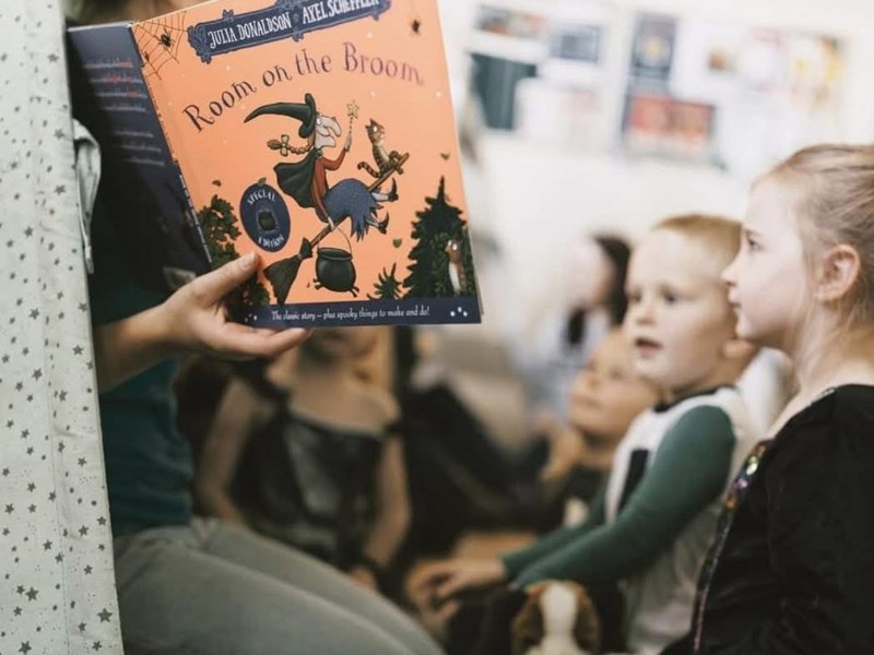 A person reads the children's book "Room on the Broom" by Julia Donaldson and Axel Scheffler to a group of young children seated attentively. The book cover shows a witch flying on a broomstick with animals. The background includes a blurred classroom with posters.