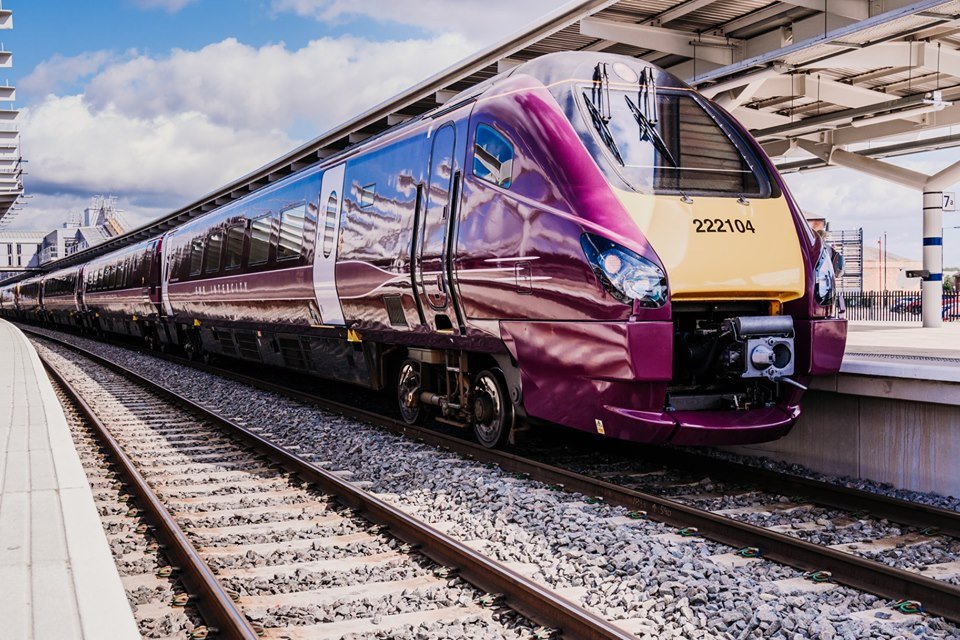 EMR Gold Award 222 - Modern maroon and cream-colored passenger train with the number 222104 parked at a station platform. The train has a sleek aerodynamic design with reflective windows, and the station features a covered roof structure with metal beams. Bright daylight and partly cloudy skies are visible in the background.