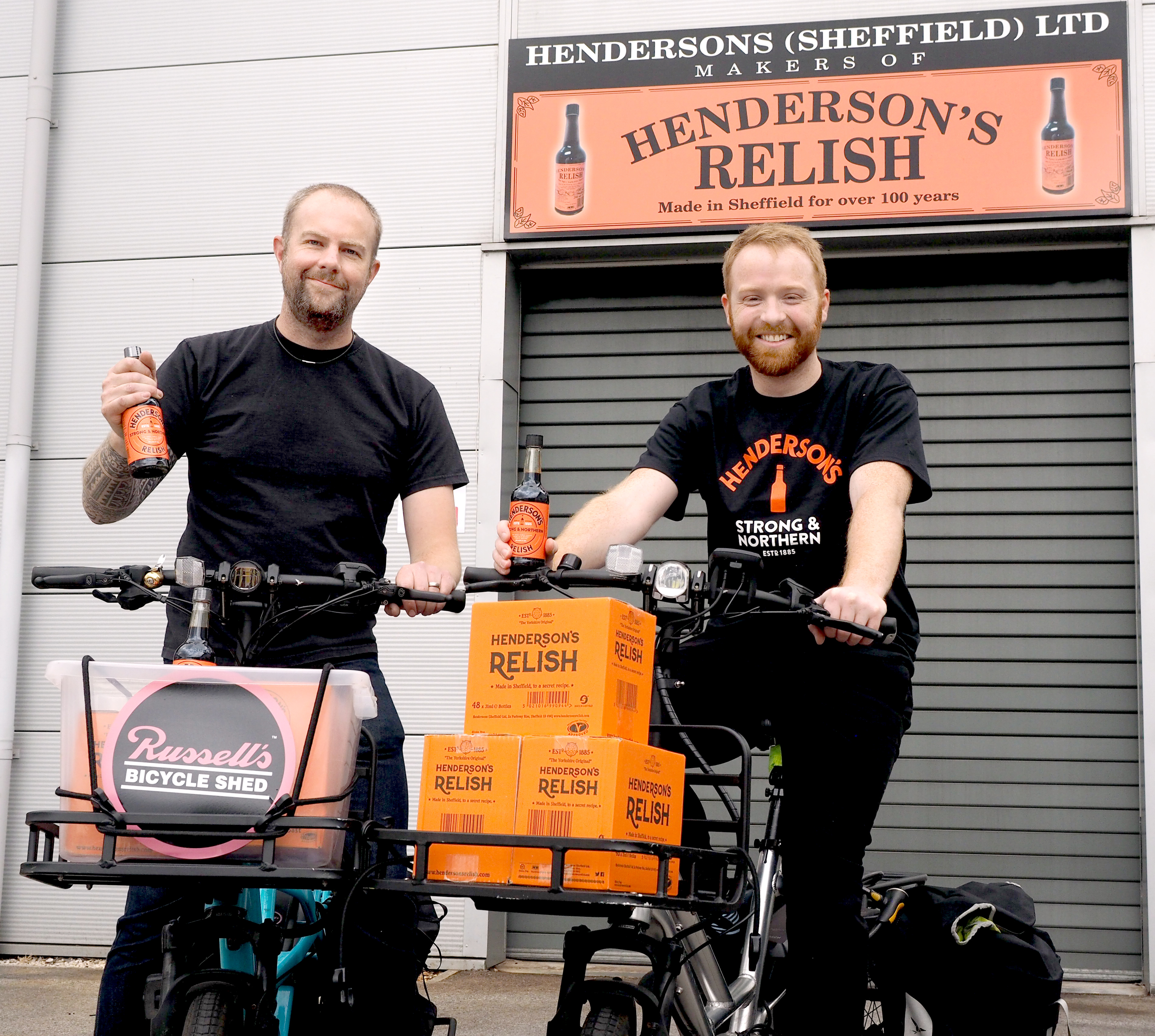 Two people standing with bicycles in front of a building featuring a sign that reads “Hendersons (Sheffield) Ltd Makers of Henderson’s Relish – Made in Sheffield for over 100 years.” Both bicycles have baskets filled with bright orange boxes labeled “Henderson’s Relish.” One person holds a bottle of Henderson’s Relish, and the other has a bottle in the basket. A smaller sign on one bike reads “Russell’s Bicycle Shed.” The background includes a grey roller shutter and the orange Henderson’s Relish branding.