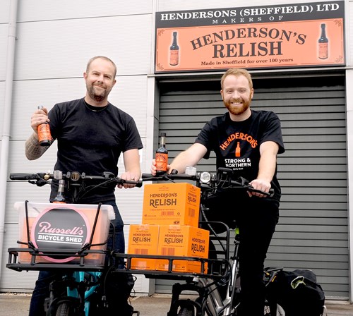Two people standing with bicycles in front of a building featuring a sign that reads “Hendersons (Sheffield) Ltd Makers of Henderson’s Relish – Made in Sheffield for over 100 years.” Both bicycles have baskets filled with bright orange boxes labeled “Henderson’s Relish.” One person holds a bottle of Henderson’s Relish, and the other has a bottle in the basket. A smaller sign on one bike reads “Russell’s Bicycle Shed.” The background includes a grey roller shutter and the orange Henderson’s Relish branding.