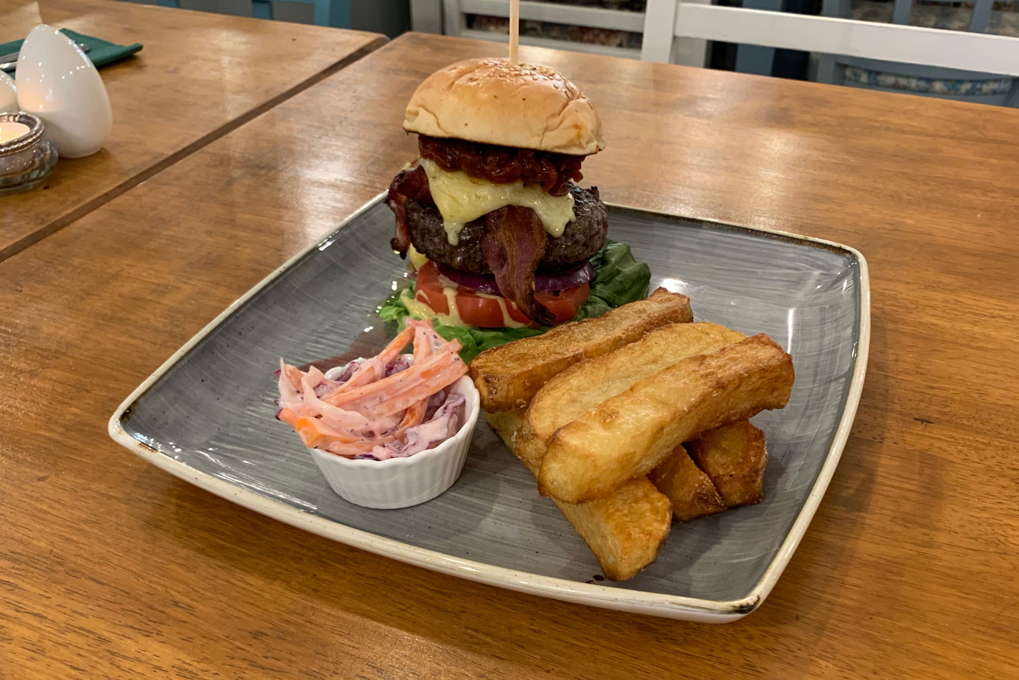 A tall burger , a stack of chunky square cut chips and a bowl of coleslaw on a square plate.