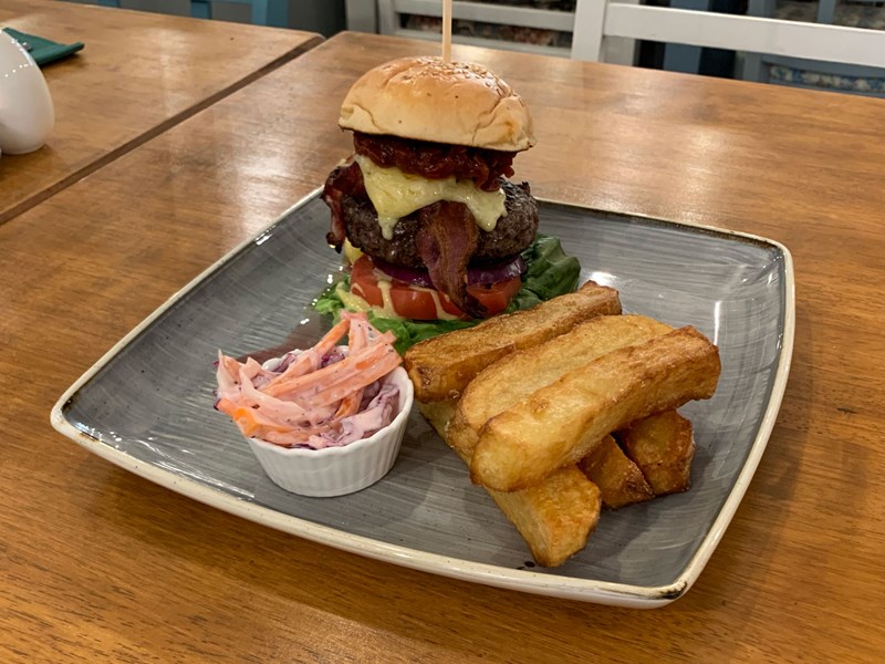 A tall burger , a stack of chunky square cut chips and a bowl of coleslaw on a square plate.