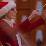 A woman in a red outfit and a Santa hat conducts an orchestra.