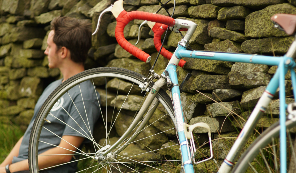 A cyclist sat leaning against a wall having a rest, out in the countryside, with his bike propped up next to him.
