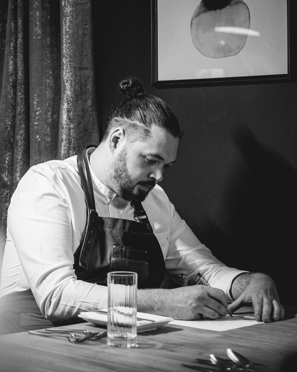 A black and white picture of a man in chef's clothing, writing at a tabel.