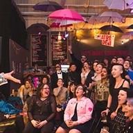 An entertainer on stage performing in front of a crowded room at Theatre Deli.