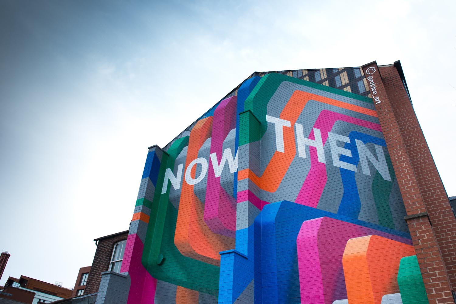 Colorful geometric mural on the side of a tall brick building featuring bold overlapping shapes in orange, pink, green, blue, and gray, with large white text reading ‘NOW THEN’ against a bright sky.