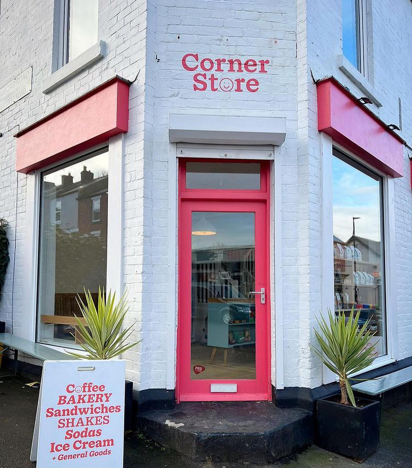 A brick building that has been painted white, with red window frames and door. There is a hand painted sign, in red, above the door that reads 'Corner Store'. The letter 'o' in the word Store is a smiley face.