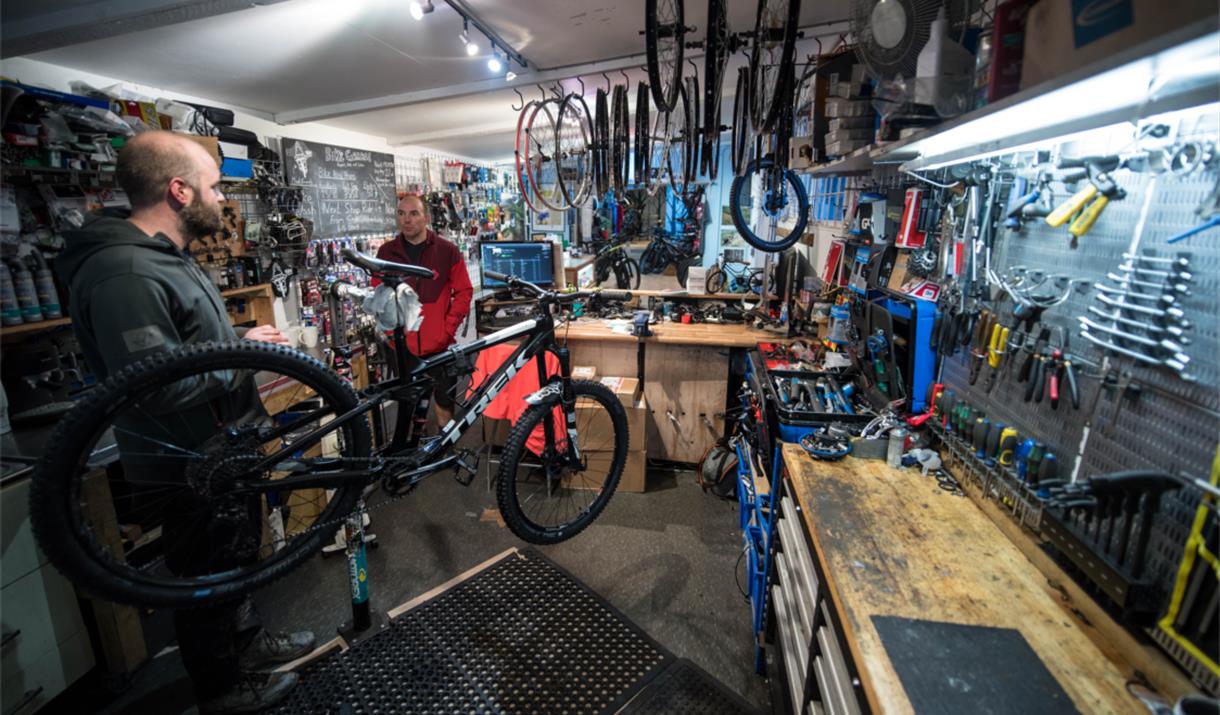 A photograph of the interior of The Bike Garage with the walls covered in tools and spare parts. There are two men working on bikes.