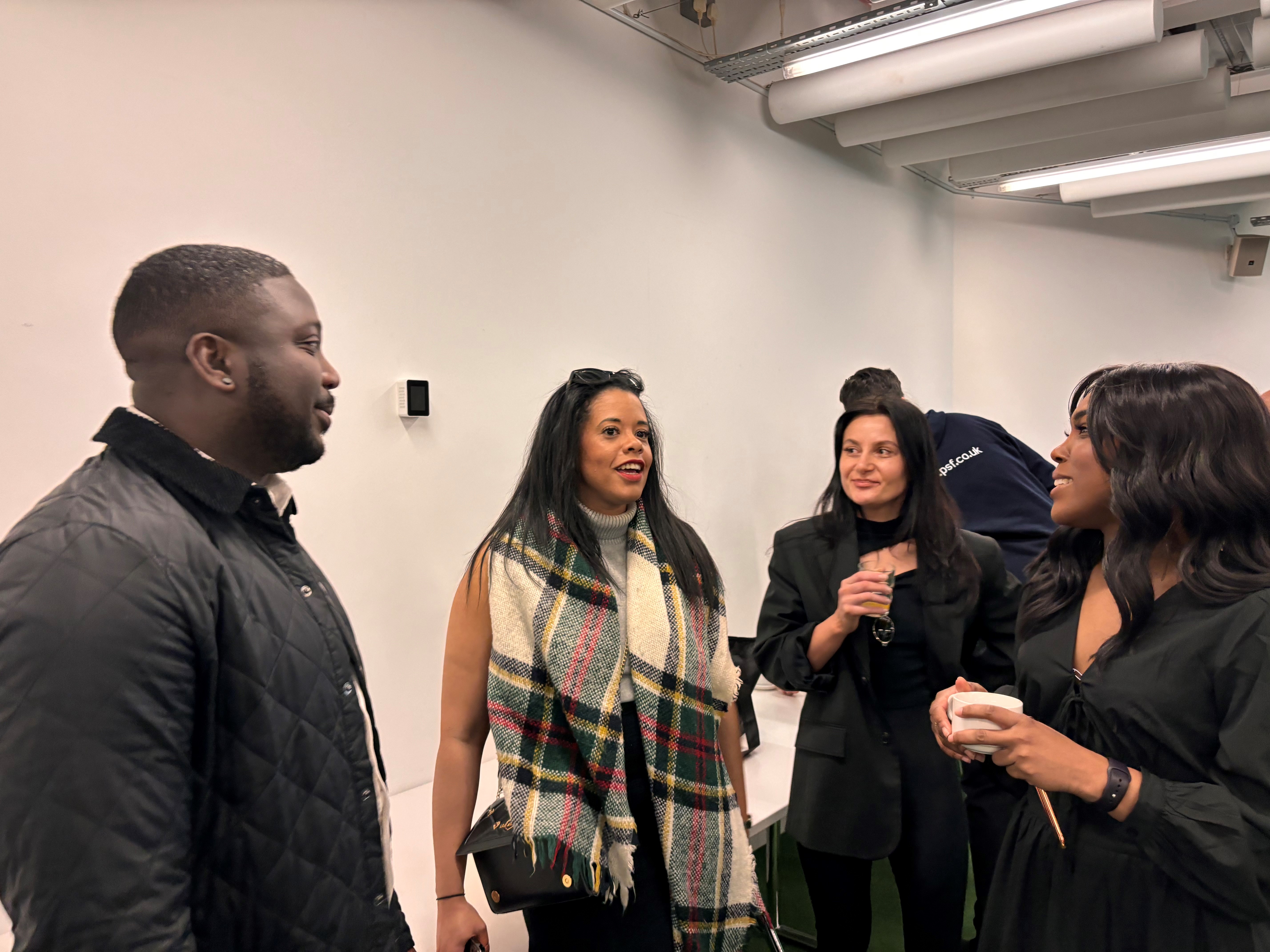 A small group of people standing indoors in conversation. The setting appears to be a modern room with white walls and exposed ceiling fixtures. One person is wearing a quilted black jacket, another has a large plaid scarf draped over their shoulders, and others are dressed in dark clothing. A white bench is visible along the wall in the background.