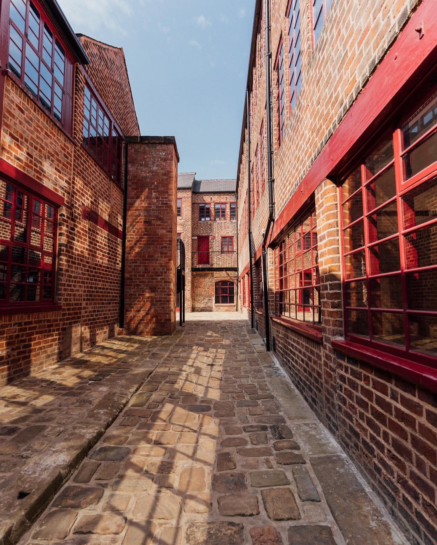  A shot of Leah's Yard, in the sunshine, in the centre of Sheffield. A cobbled path is lined on both sides by renovated red brick industrial buildings.