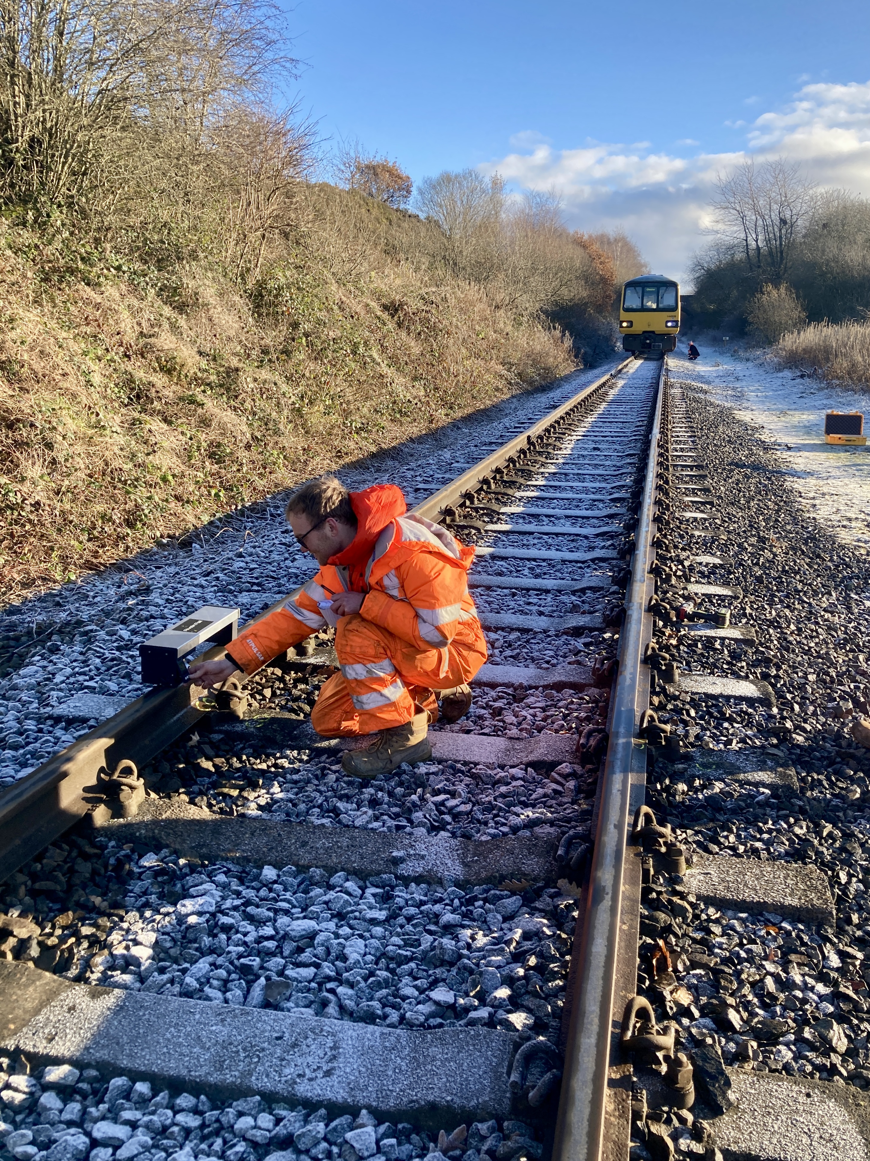 Railway maintenance worker in bright orange high-visibility gear kneeling on a frosty track, adjusting equipment attached to the rail. A train is visible in the distance on the same track, surrounded by trees and bushes under a clear blue sky.
