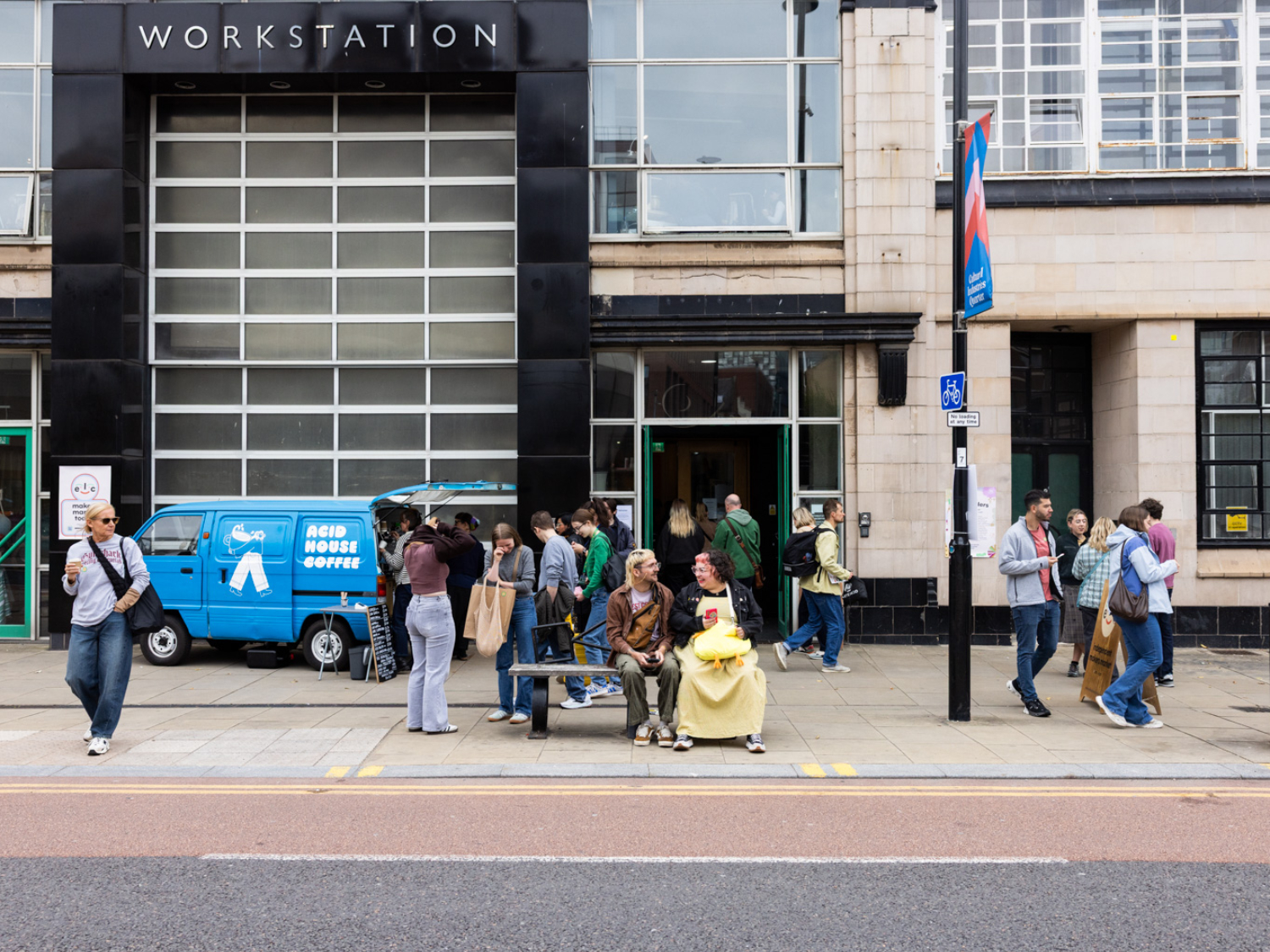 Exterior of a building labeled "WORKSTATION" with large glass windows and a black facade. A blue van marked "ACID HOUSE COFFEE" is parked in front, with people gathered around it, some in line. Others walk along the sidewalk or sit on a bench near the entrance, creating a lively urban scene.