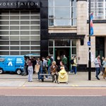 Exterior of a building labeled "WORKSTATION" with large glass windows and a black facade. A blue van marked "ACID HOUSE COFFEE" is parked in front, with people gathered around it, some in line. Others walk along the sidewalk or sit on a bench near the entrance, creating a lively urban scene.