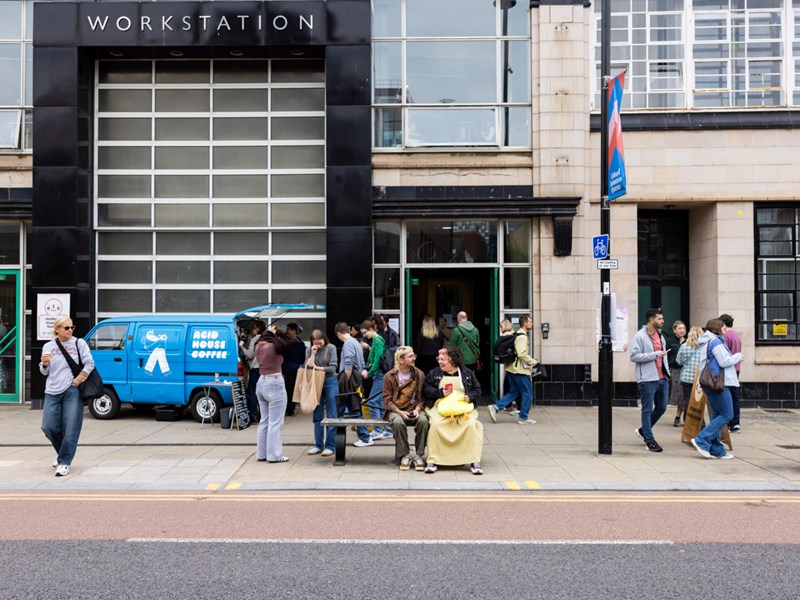 Exterior of a building labeled "WORKSTATION" with large glass windows and a black facade. A blue van marked "ACID HOUSE COFFEE" is parked in front, with people gathered around it, some in line. Others walk along the sidewalk or sit on a bench near the entrance, creating a lively urban scene.