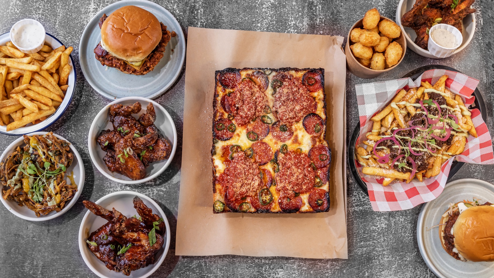 Overhead view of a food spread on a textured gray surface featuring a rectangular deep-dish pizza topped with pepperoni and cheese in the center. Surrounding dishes include burgers in buns, golden fries, crispy chicken wings, loaded fries with toppings, fried bites in a bowl, and dipping sauces. The arrangement showcases a variety of hearty comfort foods with vibrant colors and textures.