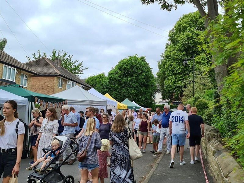 A crowd of people walking along the Nether Edge Farmers' Market. There are stalls situated in front of houses to the left, while the people walk alongside it to the right.